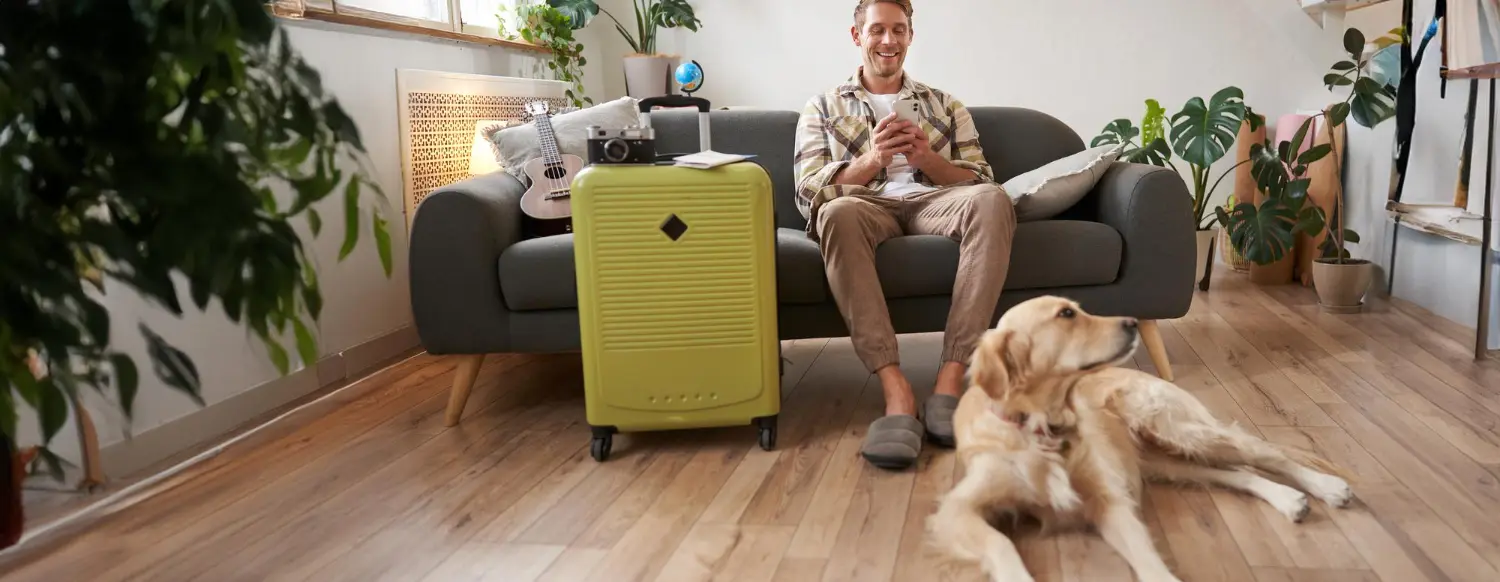 Man sitting on a sofa with a travel suitcase beside him and his golden retriever resting on the wooden floor, ready to travel abroad.